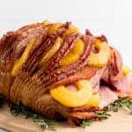 A spiral-cut baked ham garnished with caramelized pineapple rings is served on a wooden cutting board, with sprigs of fresh rosemary surrounding the baked ham.