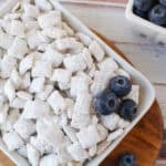 A white dish filled with Blueberry Muddy Buddies, coated in powdered sugar and topped with fresh blueberries, sits on a wooden board. A small bowl of blueberries is in the background on a light wooden surface.