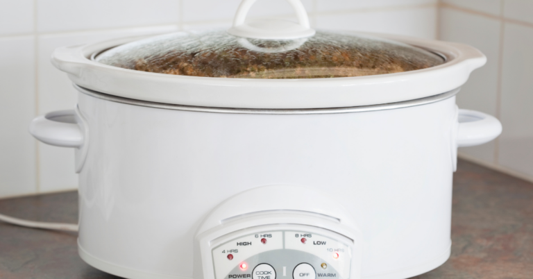 A white slow cooker with a glass lid sits on a kitchen countertop, plugged in and turned on. Steam is visible inside the lid as delicious crock pot recipes simmer. The control panel is set to a cooking mode.