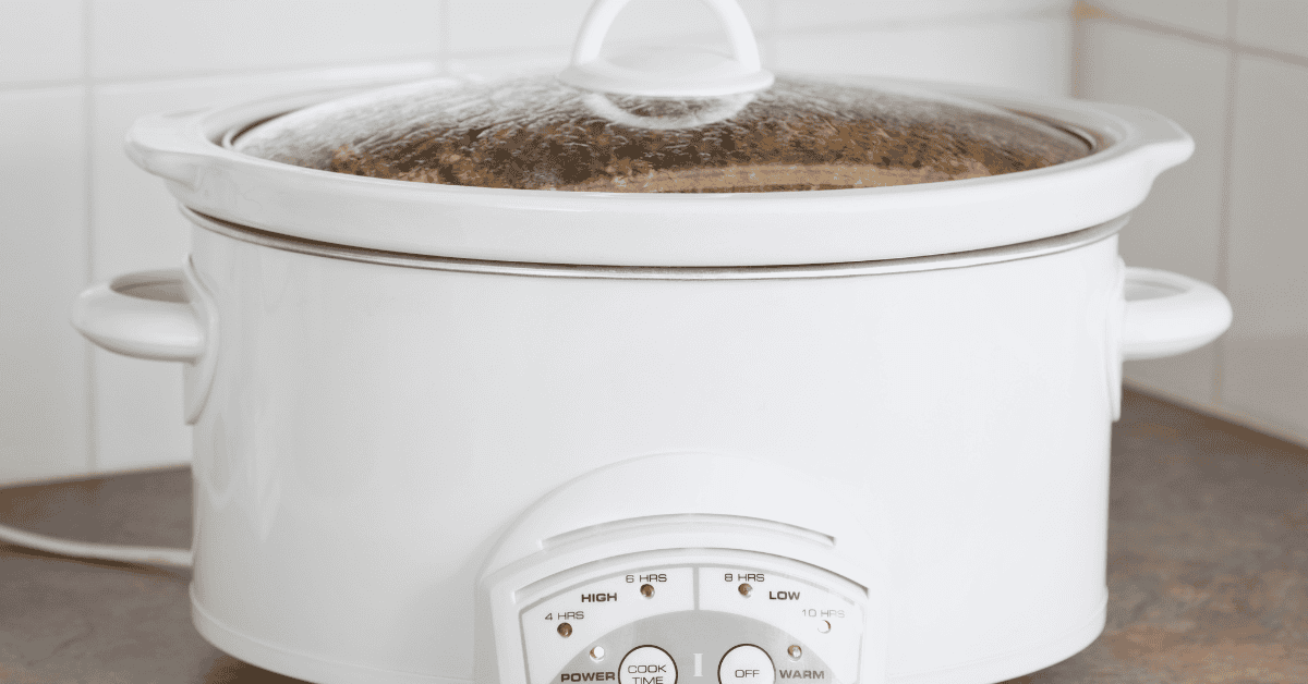 A white slow cooker with a glass lid sits on a kitchen countertop, plugged in and turned on. Steam is visible inside the lid as delicious crock pot recipes simmer. The control panel is set to a cooking mode.