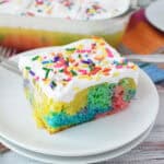 A slice of Rainbow Poke Cake topped with white frosting and rainbow sprinkles sits on a white plate, a fork beside it. The rest of the vibrant cake is in the background in a glass dish.