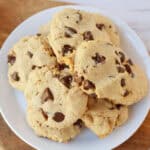 A white plate filled with chocolate chip cookies sits on a wooden surface, with golden-brown cookies scattered with chocolate chips stacked on top of each other.