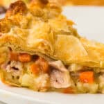 A close-up of a slice of chicken pot pie on a white plate, showcasing a golden-brown flaky crust and creamy filling with chunks of chicken, carrots, potatoes, and peas—a tempting look at this classic Chicken Pot Pie Recipe.