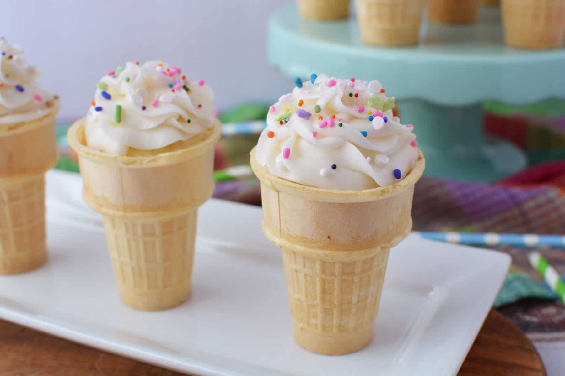 Ice Cream Cone Cupcakes with swirled white frosting and colorful sprinkles are arranged on a white plate, while more cupcakes stand elegantly in the background.
