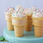 Six Ice Cream Cone Cupcakes, topped with swirled white frosting and colorful sprinkles, are displayed on a light blue cake stand.