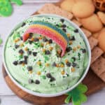 A bowl of Leprechaun Booty Dip, light green and topped with chocolate chips, sprinkles, gold pearls, and a rainbow candy. Vanilla wafers, graham crackers, and pretzels surround it on a wooden board with shamrock decorations nearby.