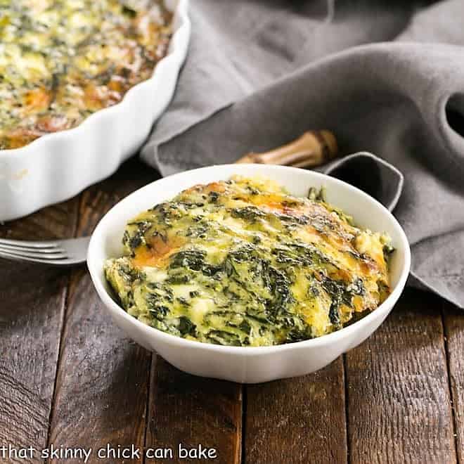 A white bowl filled with creamy, baked spinach casserole sits on a wooden table, next to a grey napkin and a fork. The casserole is golden brown on top with visible spinach and cheese.