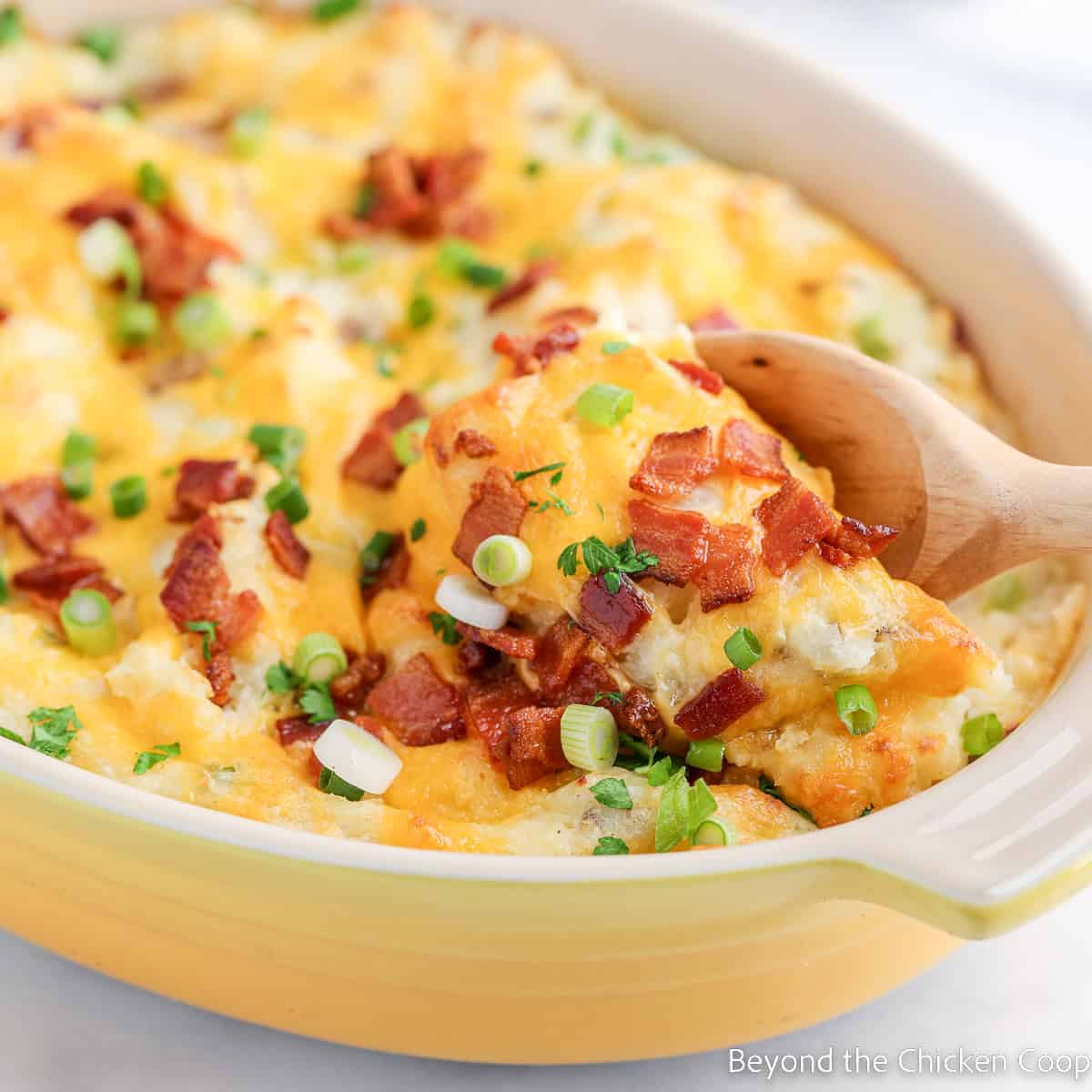 A wooden spoon lifts a serving of cheesy potato casserole topped with crispy bacon bits and chopped green onions from a yellow baking dish.