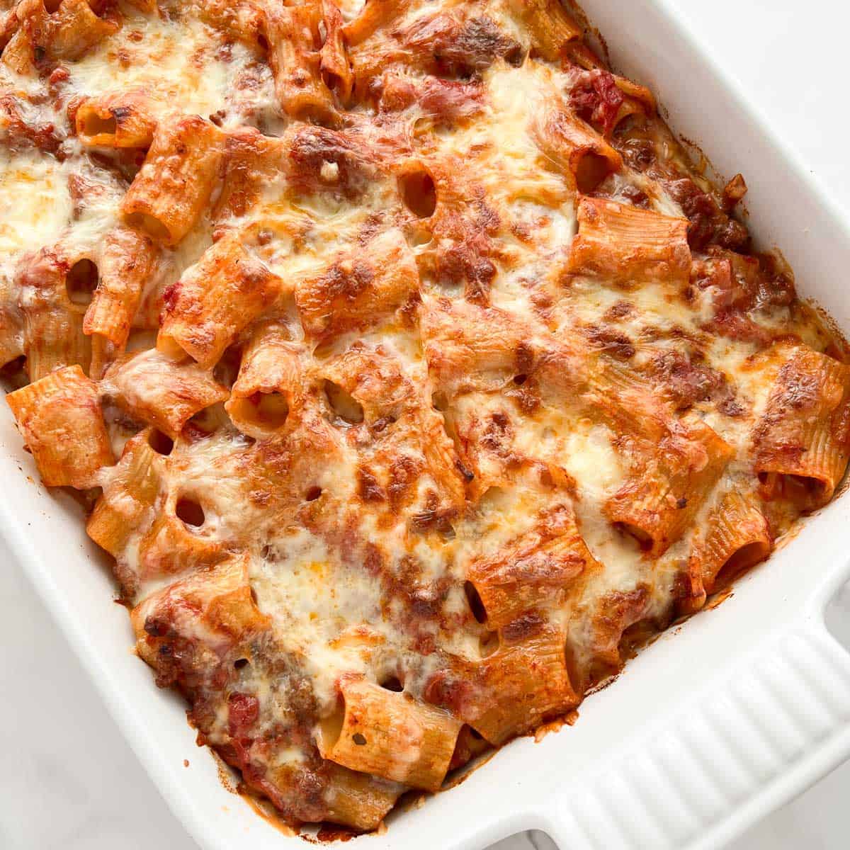 A close-up of a baked pasta dish in a white casserole dish, featuring rigatoni noodles mixed with tomato sauce and topped with melted, golden-brown cheese.