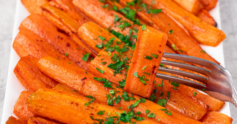 A close-up of Baked Carrots with Brown Sugar, garnished with chopped parsley on a white plate. A fork holds one perfectly glazed carrot stick above the others.