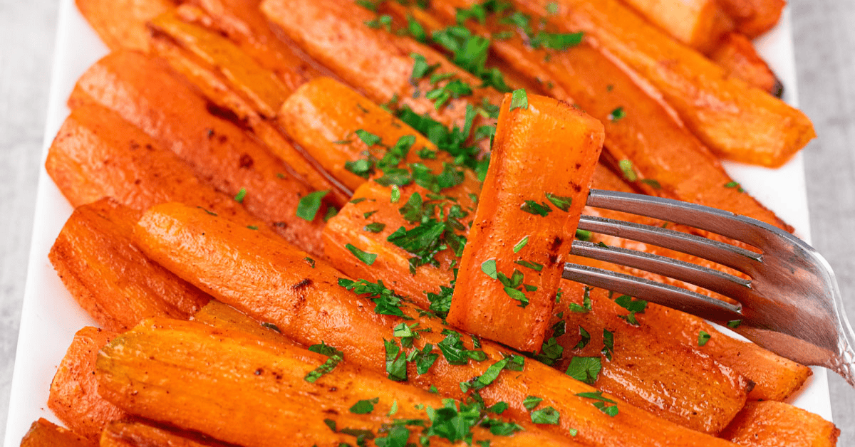 A close-up of Baked Carrots with Brown Sugar, garnished with chopped parsley on a white plate. A fork holds one perfectly glazed carrot stick above the others.