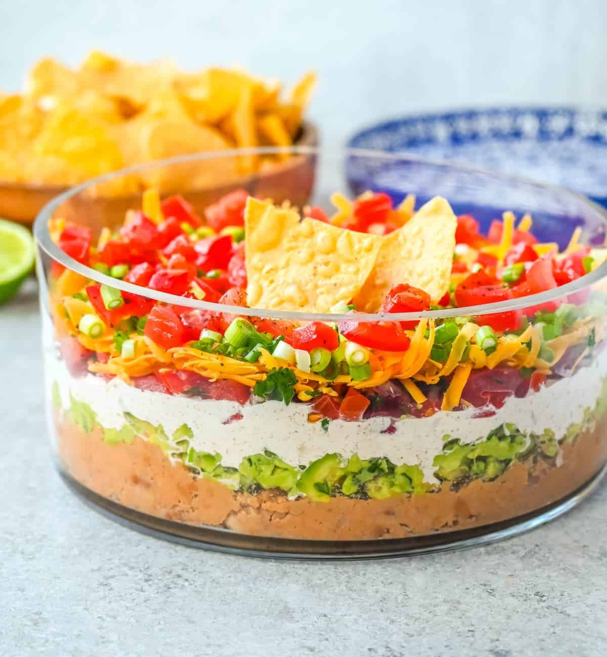 A glass bowl filled with colorful seven-layer dip, including refried beans, guacamole, sour cream, cheese, tomatoes, green onions, and topped with tortilla chips. Bowls of chips and dip are in the background.