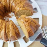 A caramel-glazed Biscoff Bundt cake topped with crushed crumbs sits on a white platter. A slice is being lifted with a spatula, and several forks are arranged beside the platter on a burlap mat.