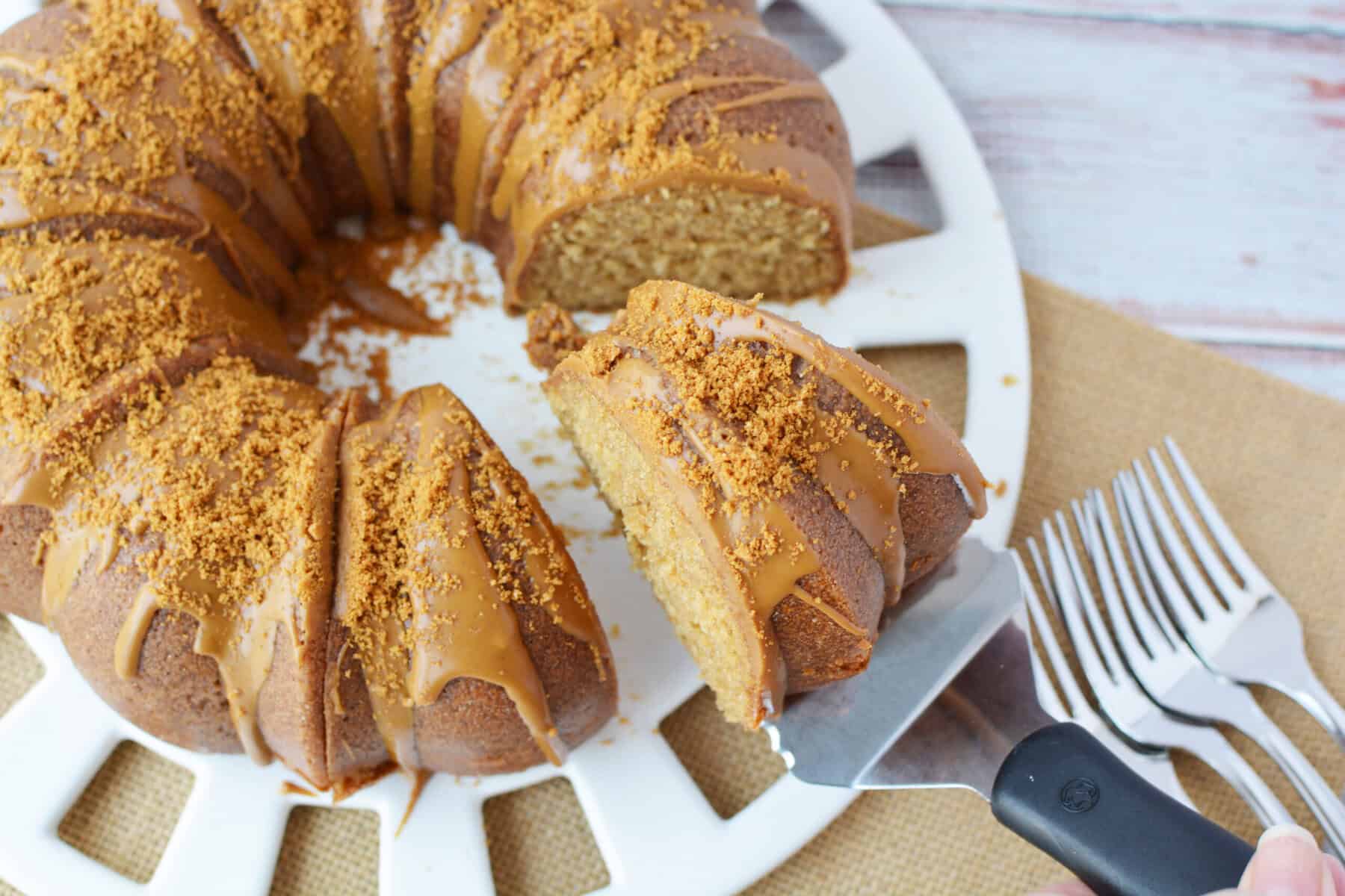 A caramel-glazed Biscoff Bundt cake topped with crushed crumbs sits on a white platter. A slice is being lifted with a spatula, and several forks are arranged beside the platter on a burlap mat.