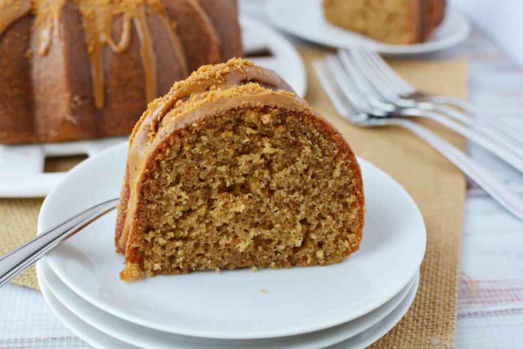 A close-up of a slice of Biscoff Bundt cake with caramel glaze on a white plate, with a fork beside it; more cake, plates, and forks are in the background.