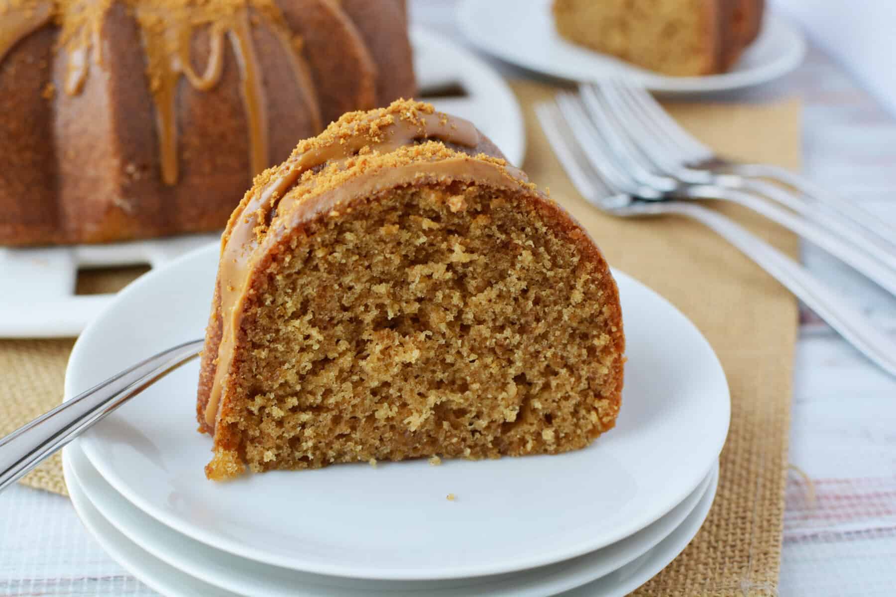 A close-up of a slice of Biscoff Bundt cake with caramel glaze on a white plate, with a fork beside it; more cake, plates, and forks are in the background.