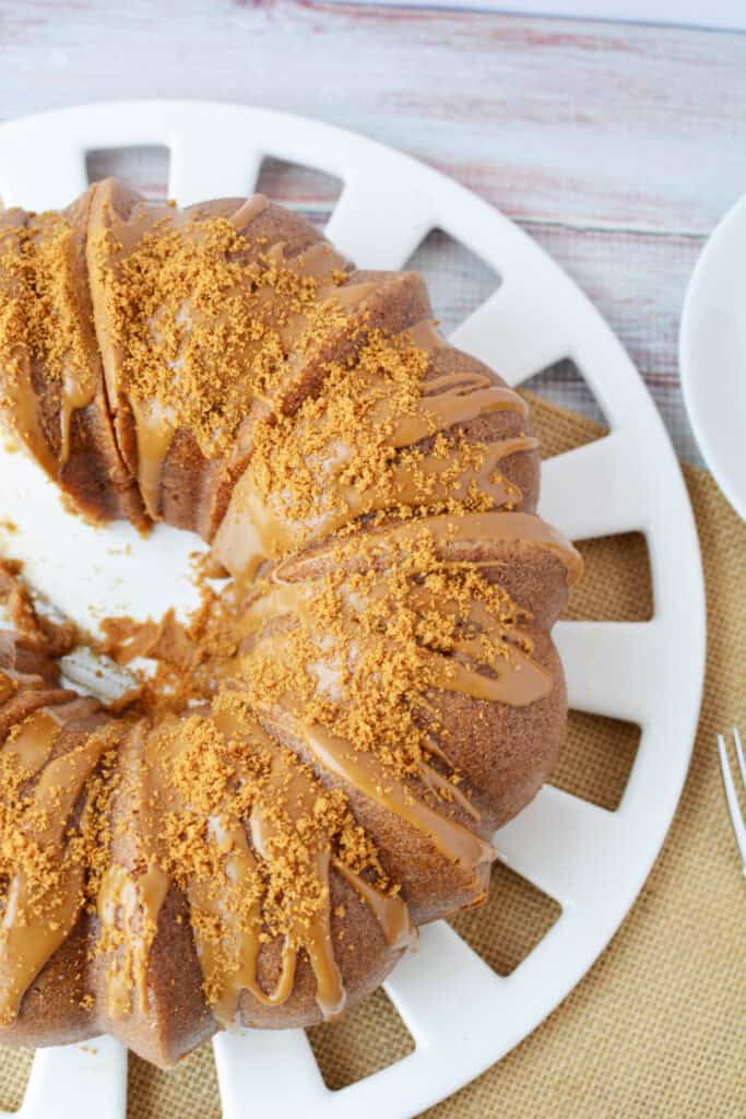 A Biscoff Bundt cake topped with caramel glaze and crushed cookie crumbs sits on a white plate, with part of the cake sliced, on a rustic wooden table. A fork and napkin are visible nearby.