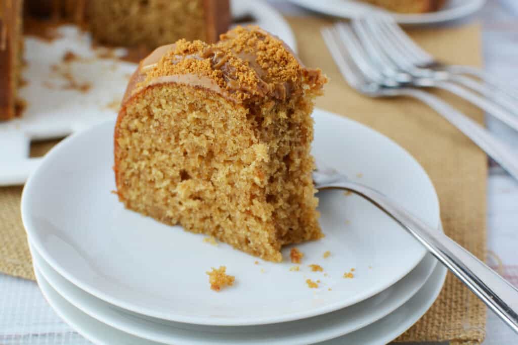 A slice of moist Biscoff Bundt cake with crumb topping sits on a white plate with a fork, stacked on other plates; blurred forks and more cake are in the background.