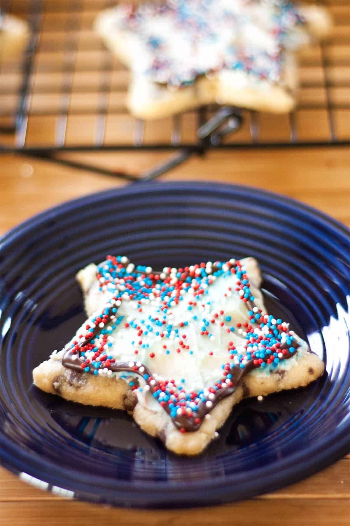 A star-shaped cookie with white icing and red, white, and blue sprinkles sits on a dark blue plate. Another similar cookie is visible in the background on a cooling rack.