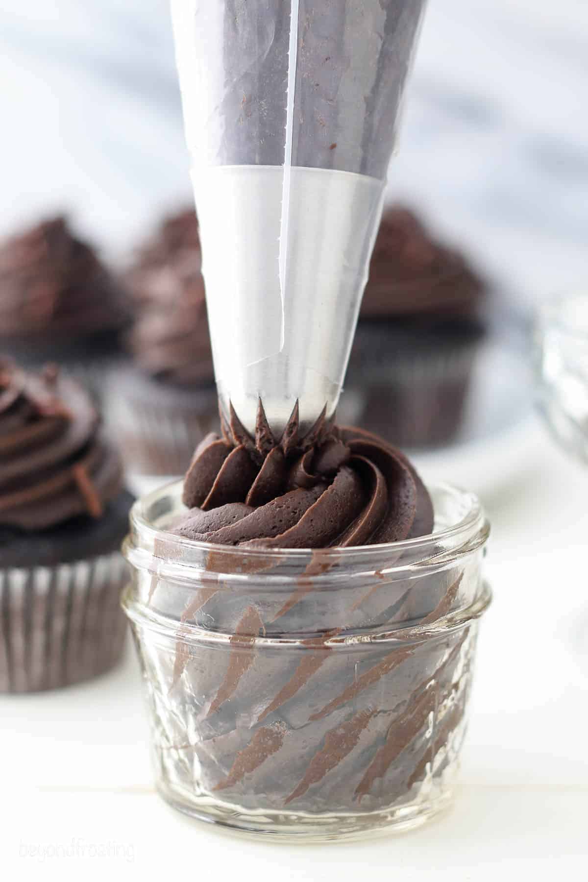 A piping bag filled with chocolate frosting is being used to decorate a small glass jar filled with chocolate cupcake, with more frosted cupcakes visible in the background.