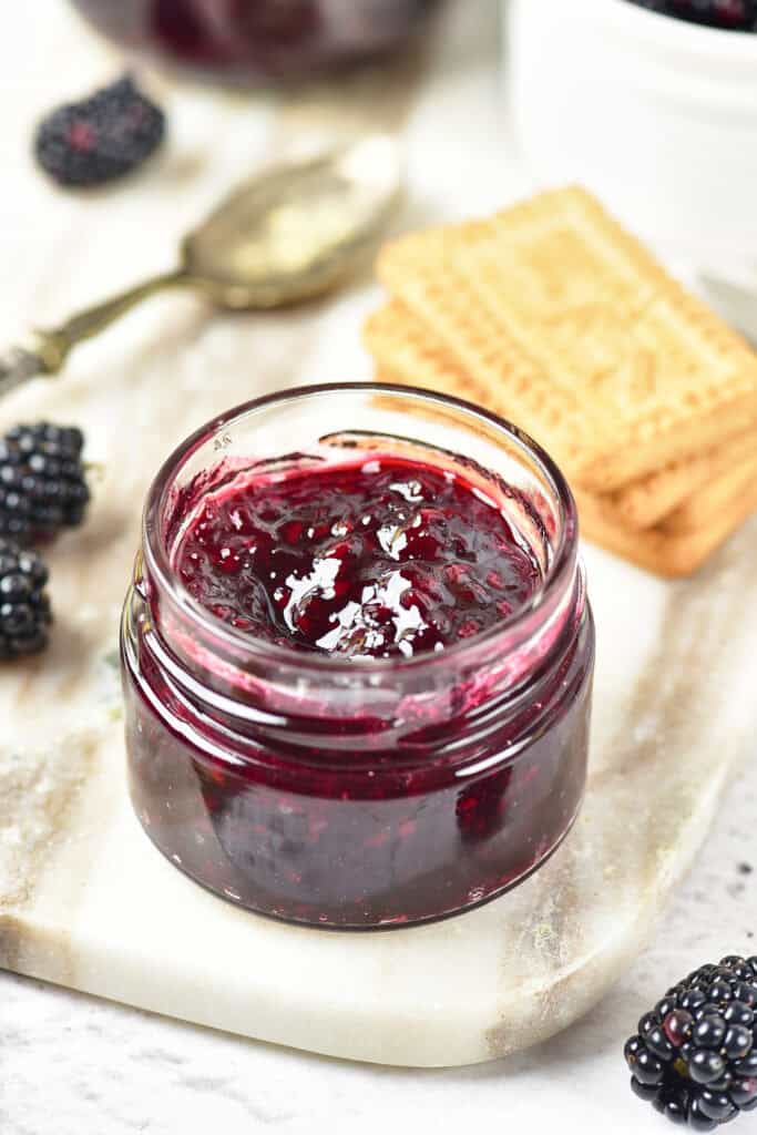 A small glass jar filled with dark blackberry jam sits on a marble board, surrounded by fresh blackberries, rectangular cookies, and a vintage spoon in the background—a perfect scene to inspire your next homemade blackberry jam recipe.