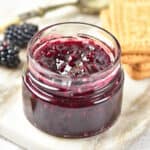 A small glass jar of blackberry jam, made from a delightful blackberry jam recipe, sits on a marble board, with fresh blackberries and rectangular biscuits nearby.