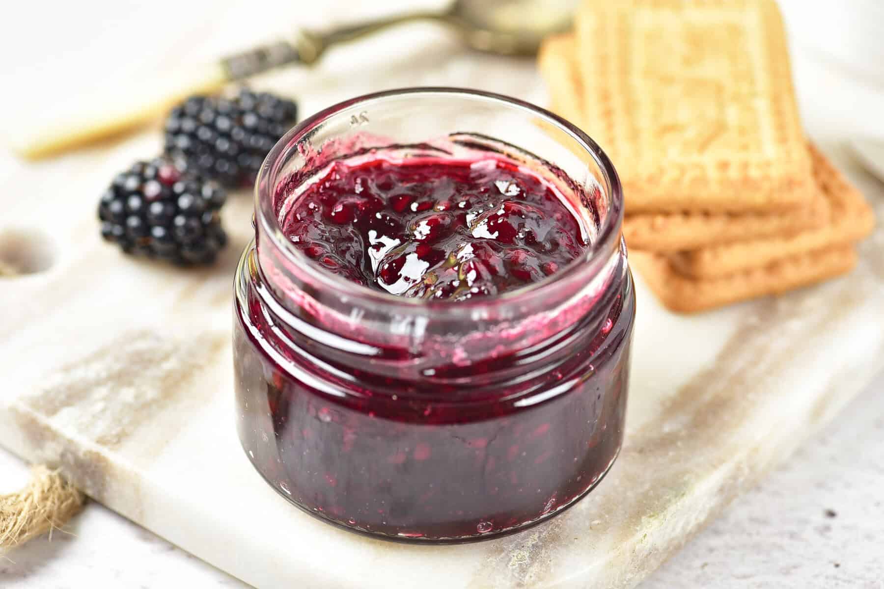 A small glass jar of blackberry jam, made from a delightful blackberry jam recipe, sits on a marble board, with fresh blackberries and rectangular biscuits nearby.