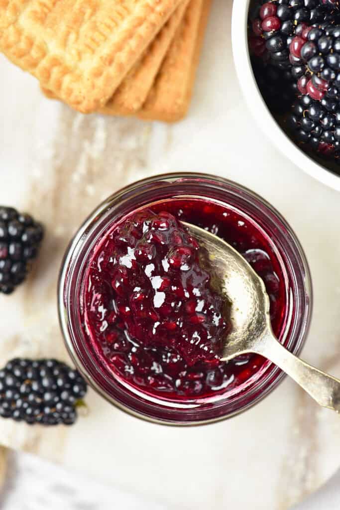 A jar of blackberry jam with a spoon, surrounded by fresh blackberries, a bowl of berries, and square biscuits on a light surface—perfect inspiration for your next homemade blackberry jam recipe.