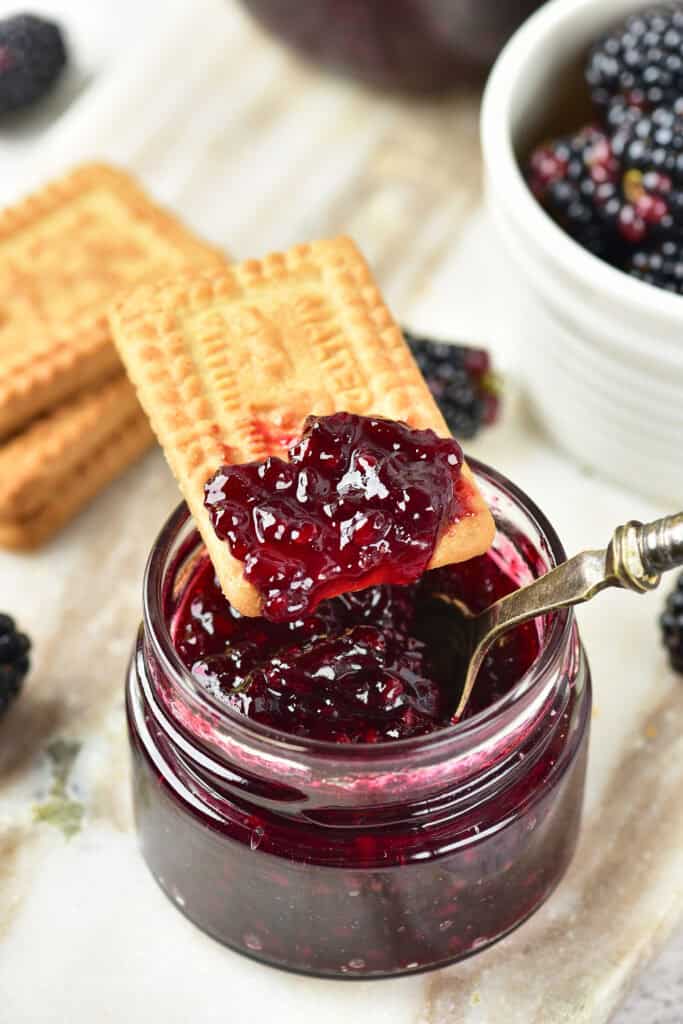 A rectangular biscuit dipped in a glass jar of blackberry jam, made from a delightful blackberry jam recipe, with a spoon inside the jar and fresh blackberries and more biscuits in the background.