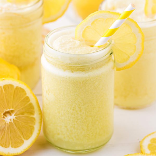 A glass jar filled with a frothy yellow lemon drink, garnished with a lemon slice and a yellow-and-white striped straw. Fresh lemons are scattered around on a light surface.