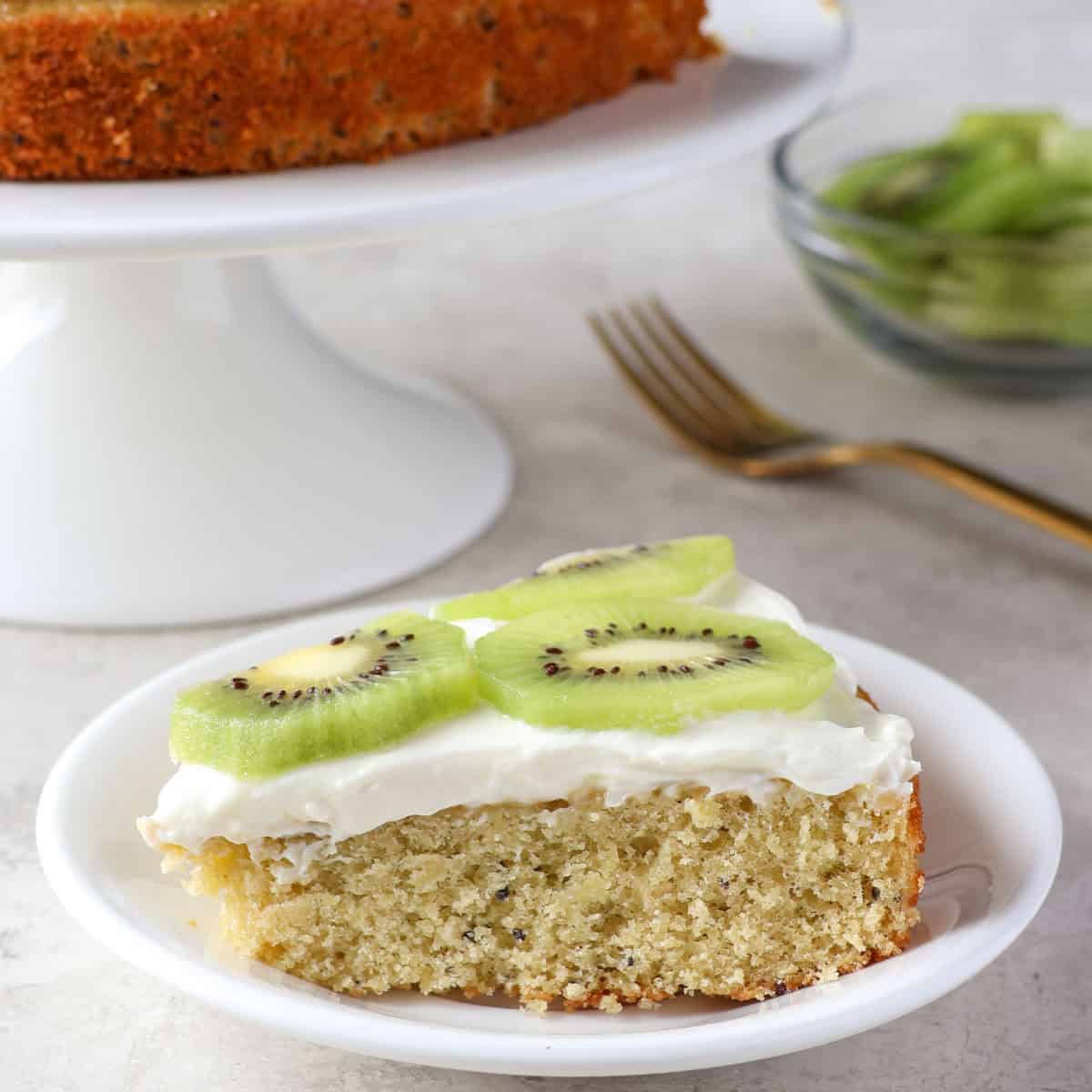 A slice of cake topped with white frosting and kiwi slices is on a white plate. The rest of the cake is on a cake stand in the background, with a fork and a bowl of sliced kiwi nearby.