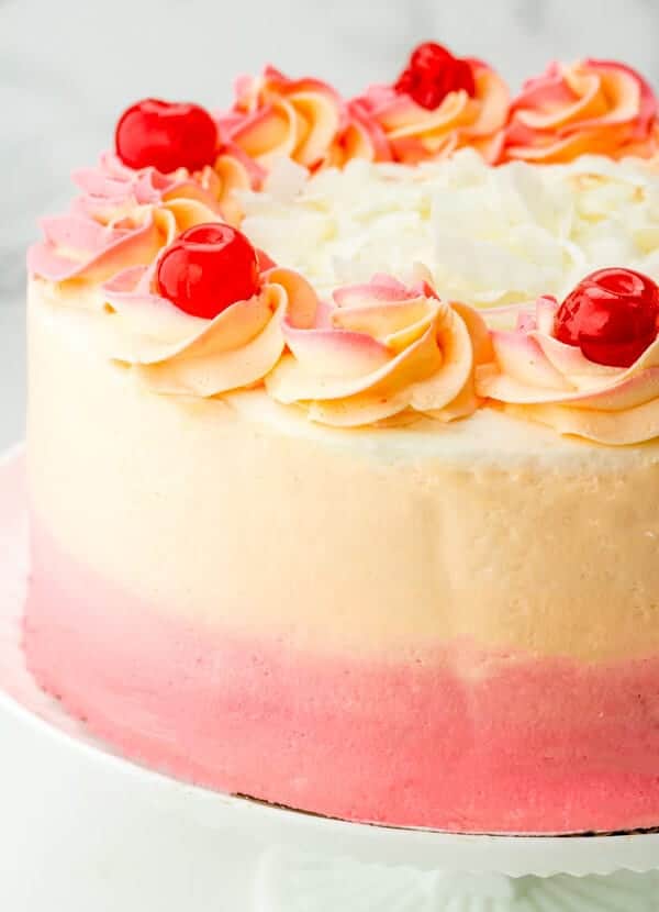 A round cake with pink and cream ombré frosting, decorated with swirls of icing, bright red cherries, and white chocolate shavings on top, displayed on a white cake stand.
