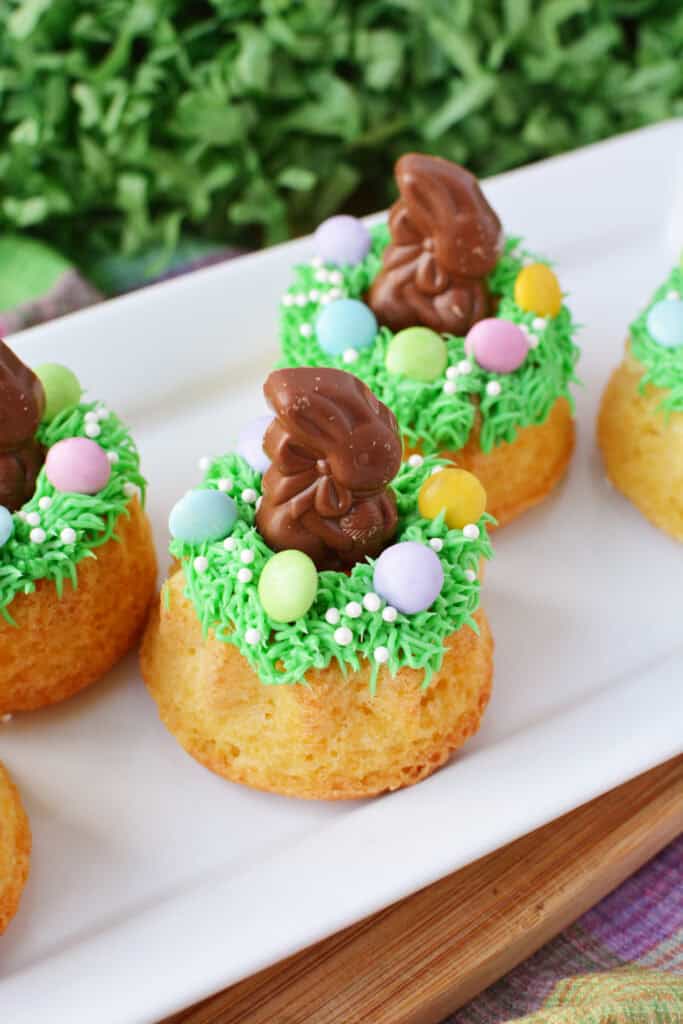Easter Mini Bundt Cakes topped with green frosting, pastel candy eggs, white sprinkles, and a chocolate bunny are arranged on a white platter with greenery in the background.
