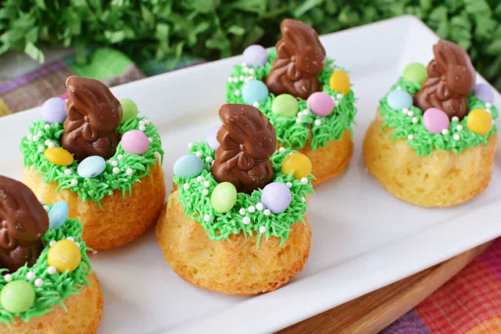 Easter Mini Bundt Cakes topped with green icing, colorful candy eggs, and small chocolate bunnies are arranged on a white platter, creating a festive spring or Easter-themed dessert display.
