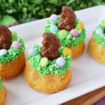 Easter Mini Bundt Cakes topped with green icing, colorful candy eggs, and small chocolate bunnies are arranged on a white platter, creating a festive spring or Easter-themed dessert display.