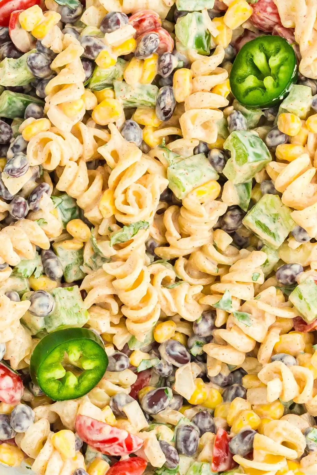 Close-up of a creamy pasta salad with rotini noodles, black beans, corn, chopped green bell peppers, cherry tomatoes, diced onions, and sliced jalapeños.