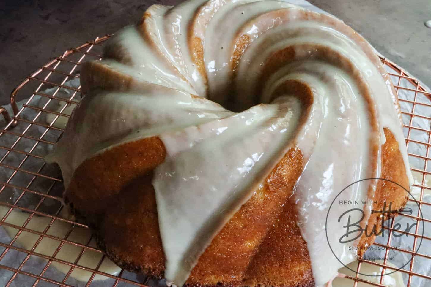 A glazed bundt cake sits on a round wire cooling rack, with icing drizzled smoothly over its ridges. The cake has a golden-brown color and appears moist and freshly baked.