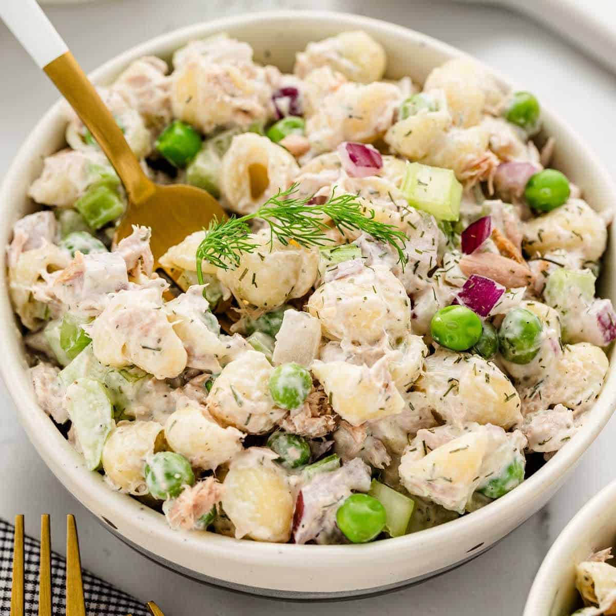 A bowl of creamy potato salad with green peas, diced celery, red onions, and fresh dill, served with a gold fork.