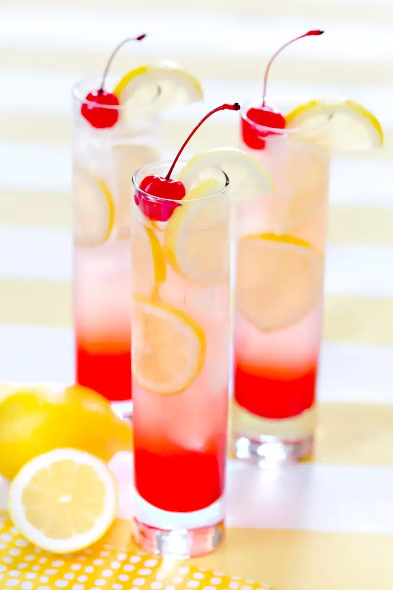 Three tall glasses of pink lemonade garnished with lemon slices and maraschino cherries, set on a yellow and white striped surface with fresh lemons nearby.