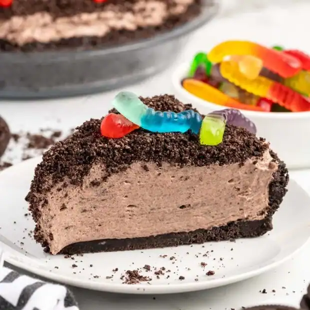 A slice of chocolate cream pie with a crushed cookie crust and topping, decorated with colorful gummy worms, sits on a white plate. In the background, there’s a bowl of more gummy worms.