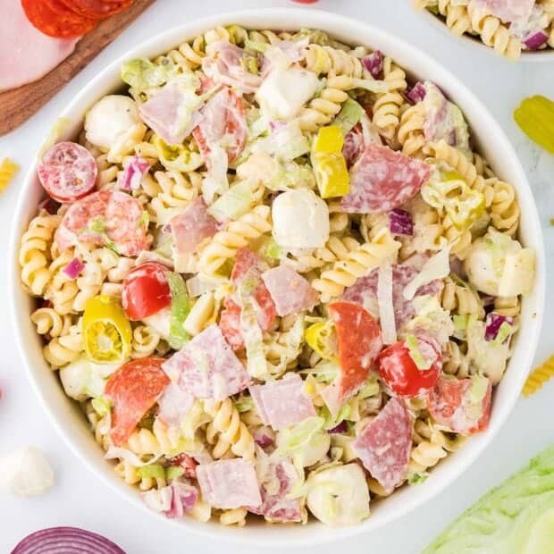 A bowl of pasta salad with rotini, sliced salami, ham, pepperoni, mozzarella balls, cherry tomatoes, red onions, lettuce, and pepperoncini, all mixed with a creamy dressing.