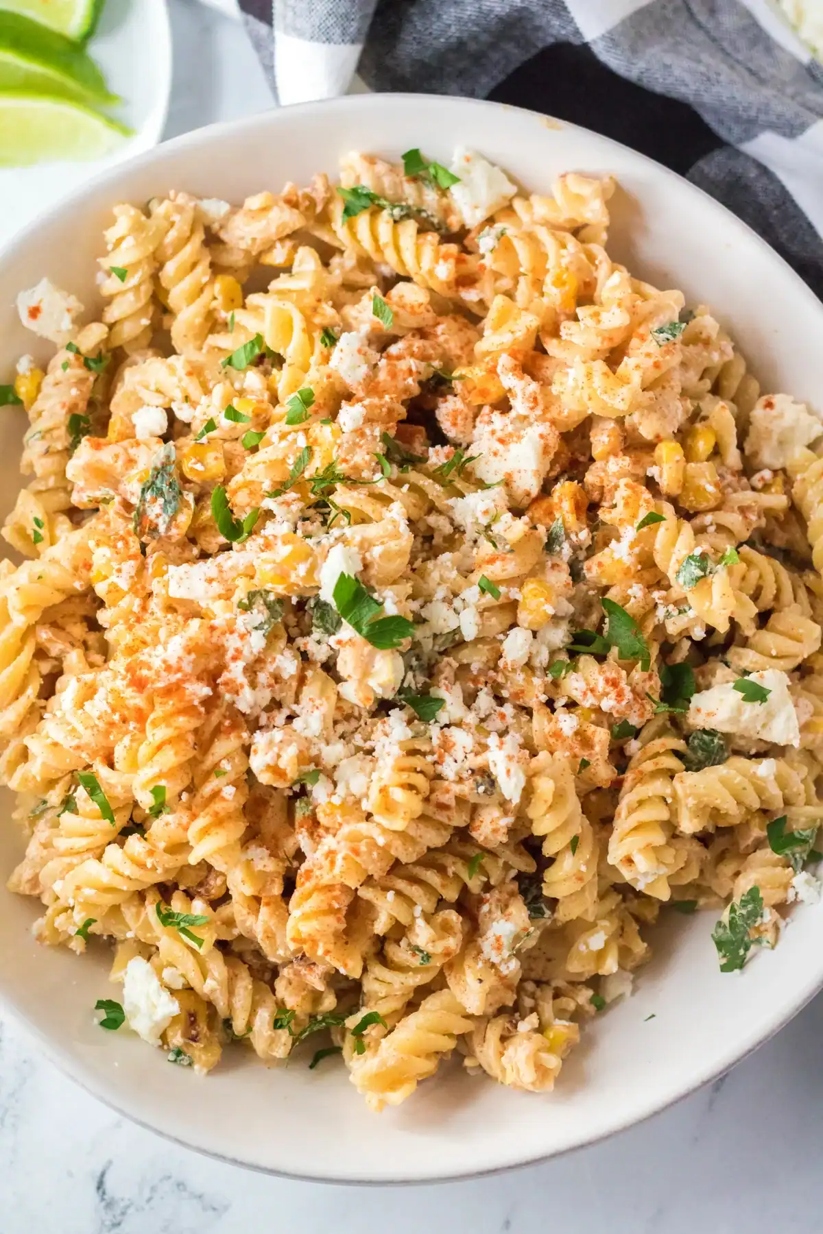 A white bowl filled with creamy rotini pasta mixed with corn, crumbled cheese, chopped herbs, and sprinkled with paprika, set on a marble surface with lime wedges in the background.