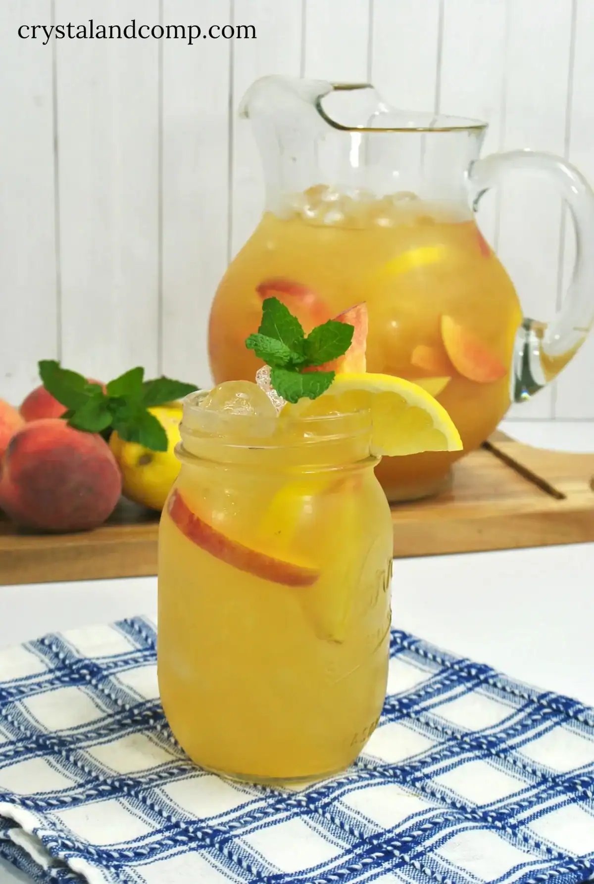 A mason jar filled with iced peach drink, garnished with a lemon slice and mint, sits on a blue-checkered cloth. A pitcher of the same drink and fresh peaches are in the background.