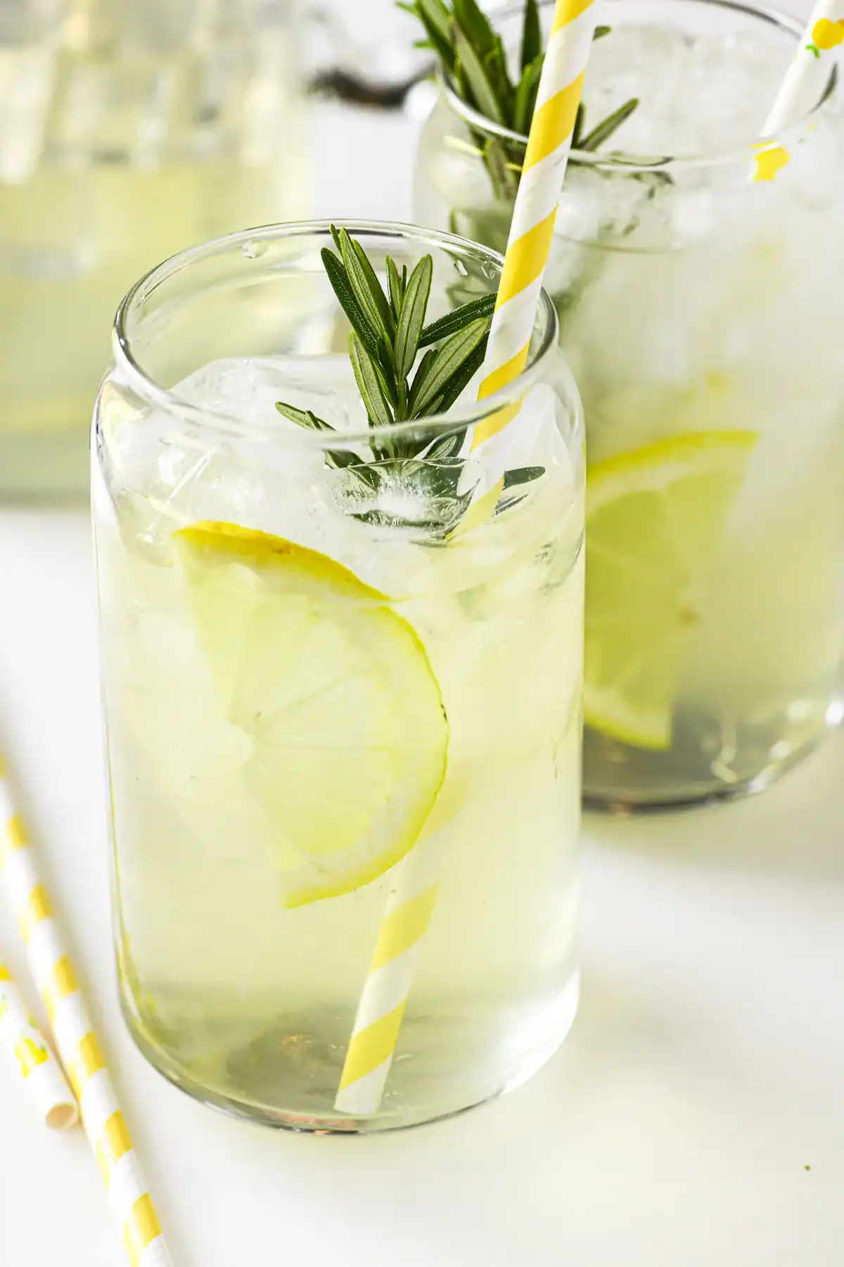 A glass of iced lemonade garnished with a lemon slice and a sprig of rosemary, served with a yellow-striped straw. Another similar glass and a pitcher are visible in the background.