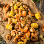 A delicious Seafood Boil featuring cooked shrimp, corn on the cob pieces, sausage slices, potatoes, and herbs is spread out on brown paper atop a rustic wooden table.