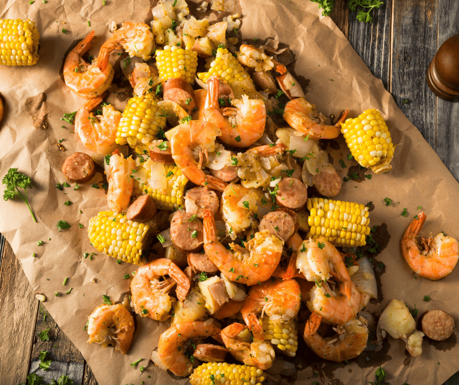 A delicious Seafood Boil featuring cooked shrimp, corn on the cob pieces, sausage slices, potatoes, and herbs is spread out on brown paper atop a rustic wooden table.