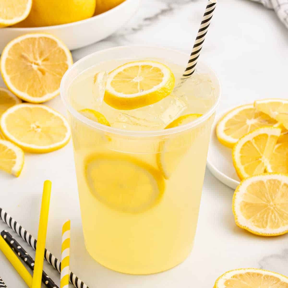 A clear plastic cup of iced lemonade with lemon slices and a black-and-white striped straw, surrounded by sliced lemons and additional straws on a white surface.