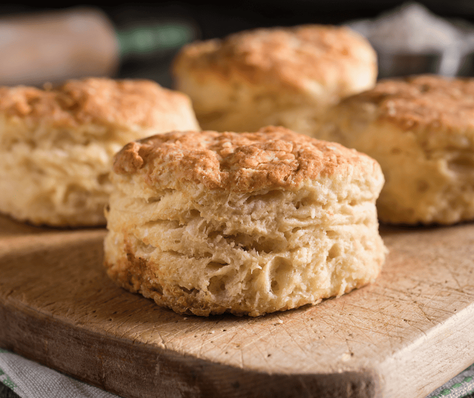 Freshly baked, golden-brown sourdough biscuits sit on a wooden cutting board, displaying a flaky, layered texture and slightly crisp crust.