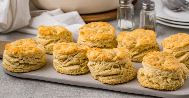 Eight golden, flaky Pancake Mix Biscuits are arranged on a rectangular tray, with a light gray countertop and kitchen items like plates, salt, and pepper shakers in the background.