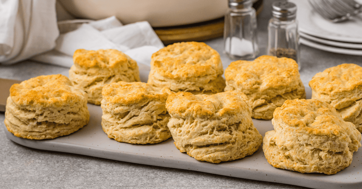 Eight golden, flaky Pancake Mix Biscuits are arranged on a rectangular tray, with a light gray countertop and kitchen items like plates, salt, and pepper shakers in the background.